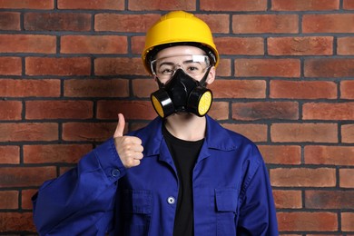 Worker in respirator, protective glasses and helmet showing thumbs up near brick wall