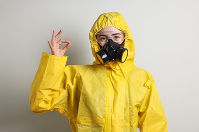 Photo of Worker in respirator, protective suit and glasses showing OK gesture on grey background