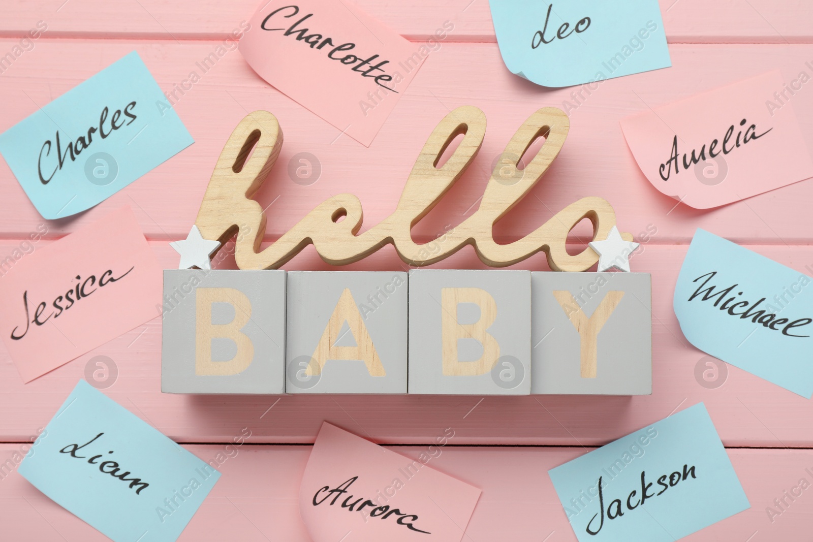Photo of Cubes with phrase Hello Baby and paper stickers with different names on pink wooden background, flat lay. Choosing baby's name