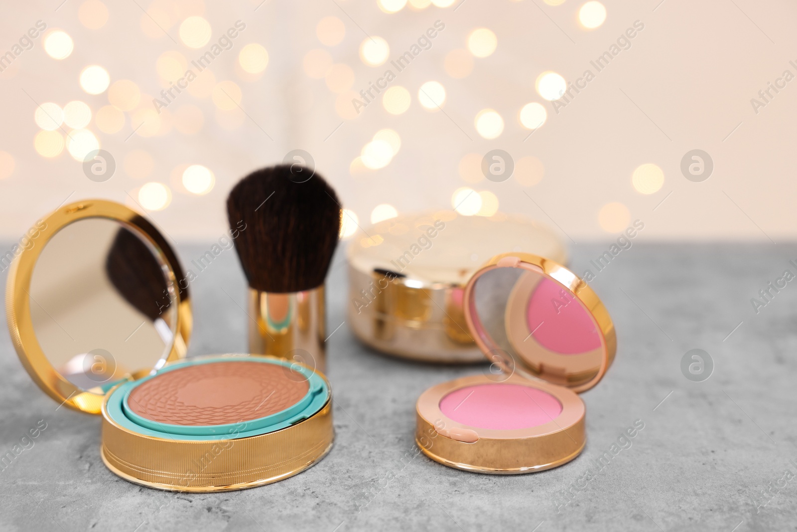 Photo of Face bronzer, blusher and other cosmetic products on grey textured table against blurred lights, closeup