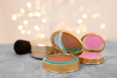 Face bronzer and other cosmetic products on grey textured table against blurred lights, closeup