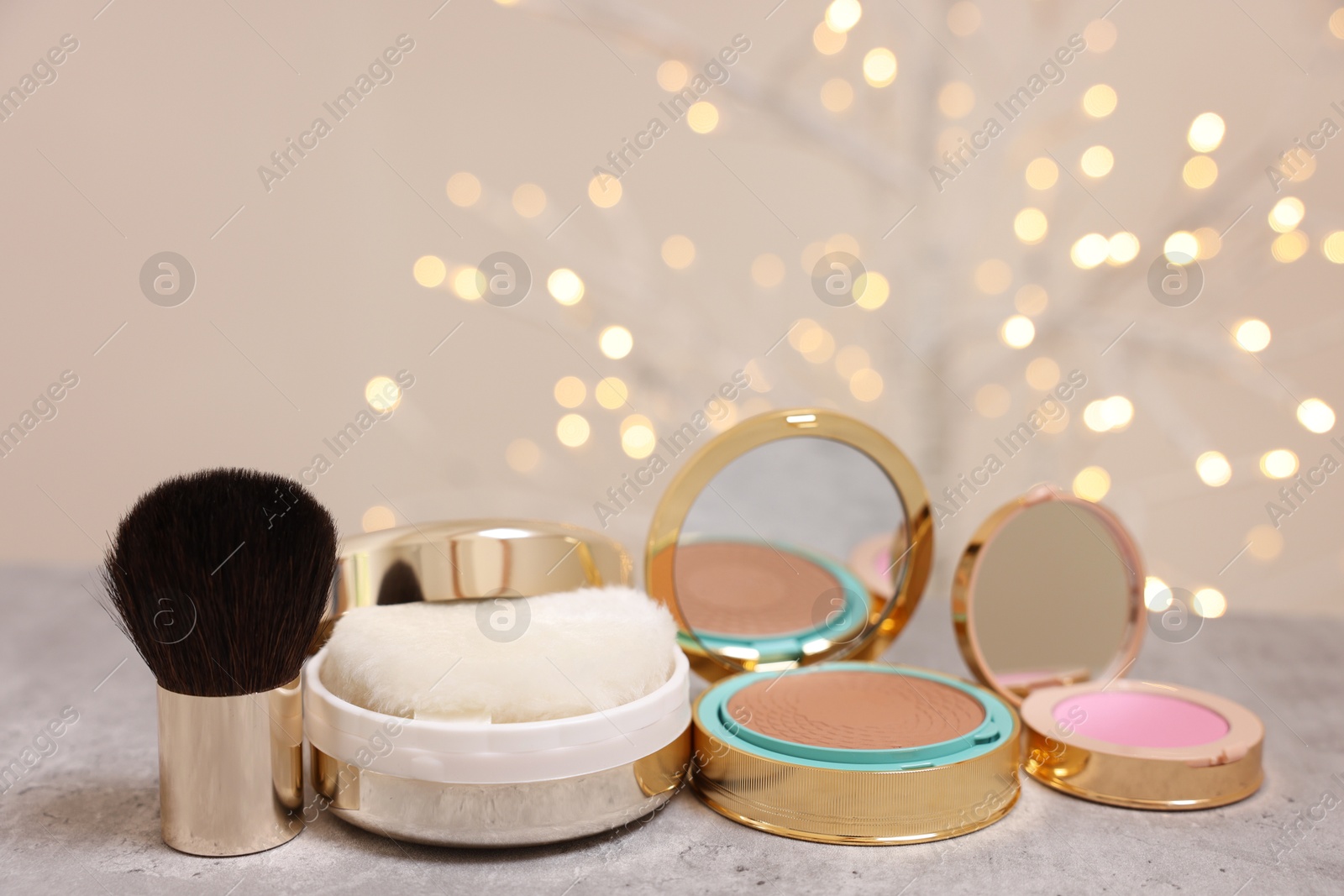 Photo of Bronzer, powder, blusher and brush on grey textured table against blurred lights, closeup