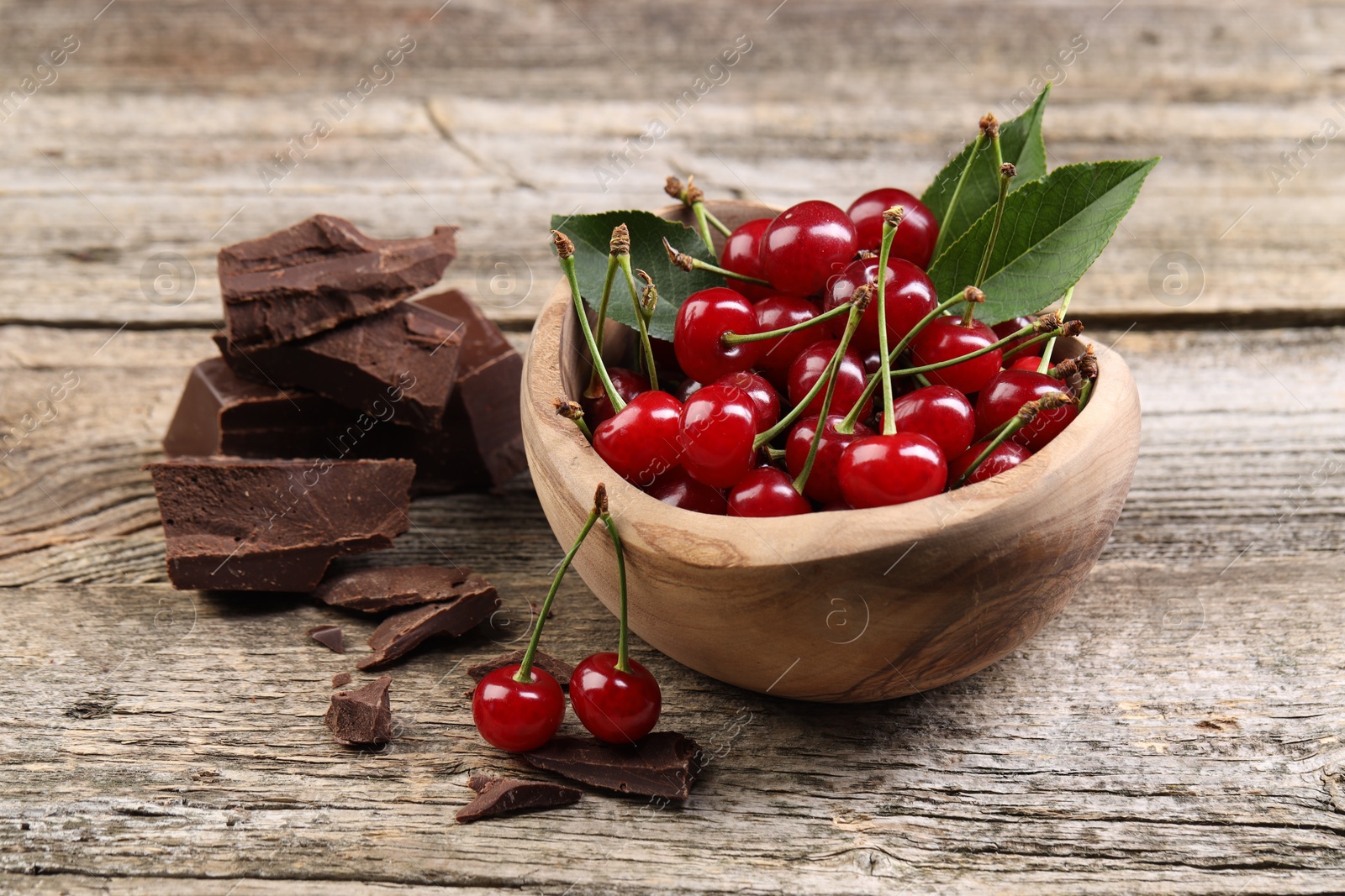 Photo of Fresh cherries with green leaves in bowl and dark chocolate on wooden table