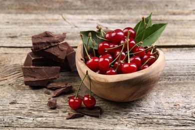 Fresh cherries with green leaves in bowl and dark chocolate on wooden table