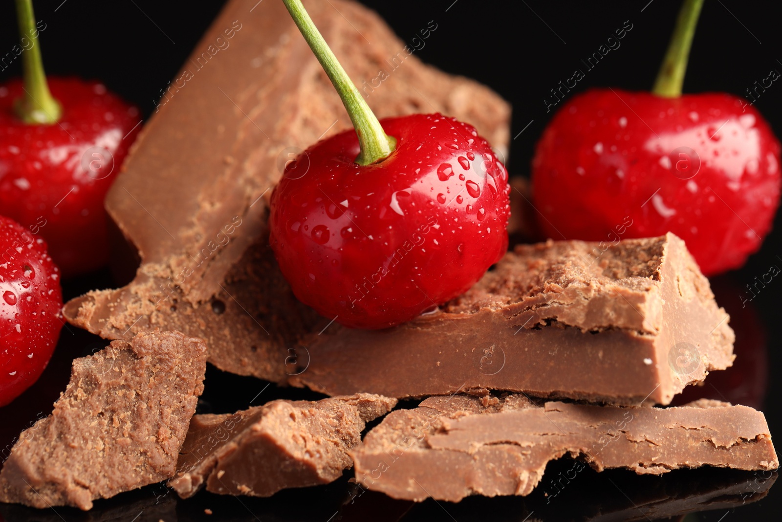 Photo of Fresh cherries with pieces of milk chocolate on black surface, closeup