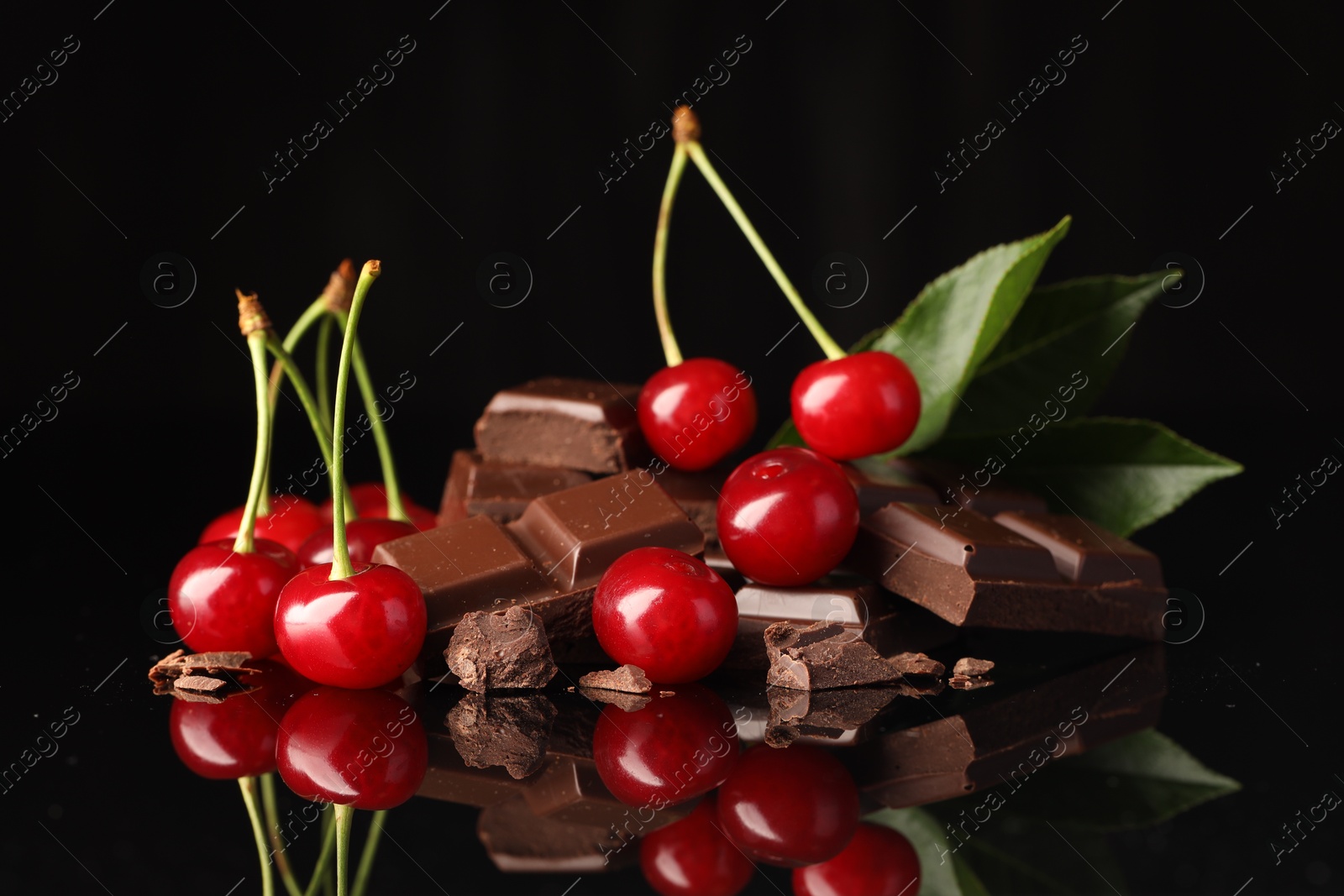 Photo of Fresh cherries with pieces of milk chocolate and green leaves on black mirror surface