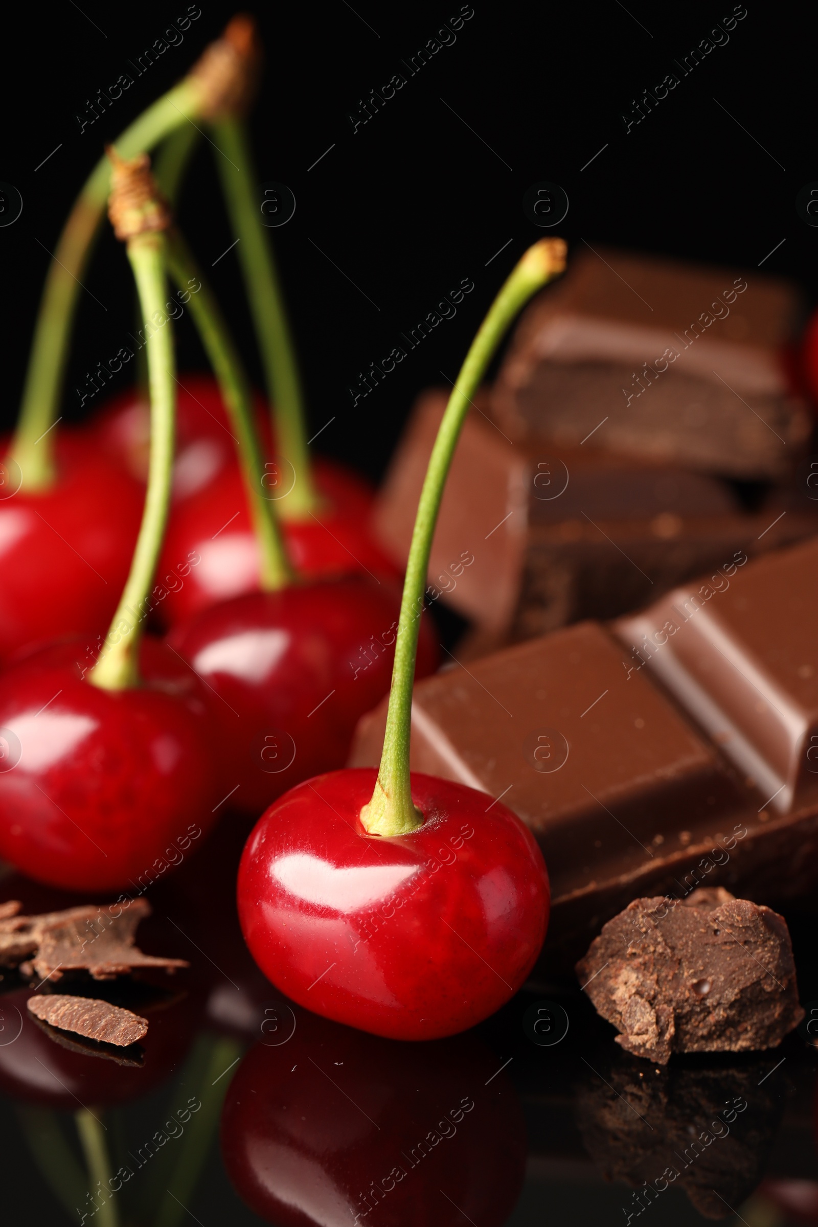 Photo of Fresh cherries with pieces of milk chocolate on black mirror surface, closeup