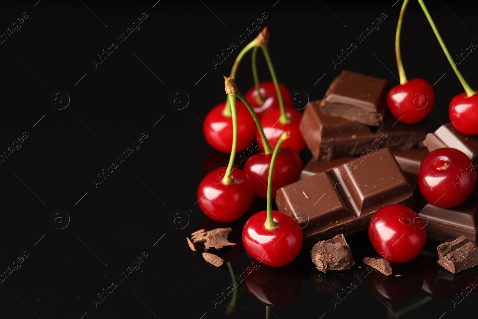 Photo of Fresh cherries with pieces of milk chocolate on black mirror surface, space for text