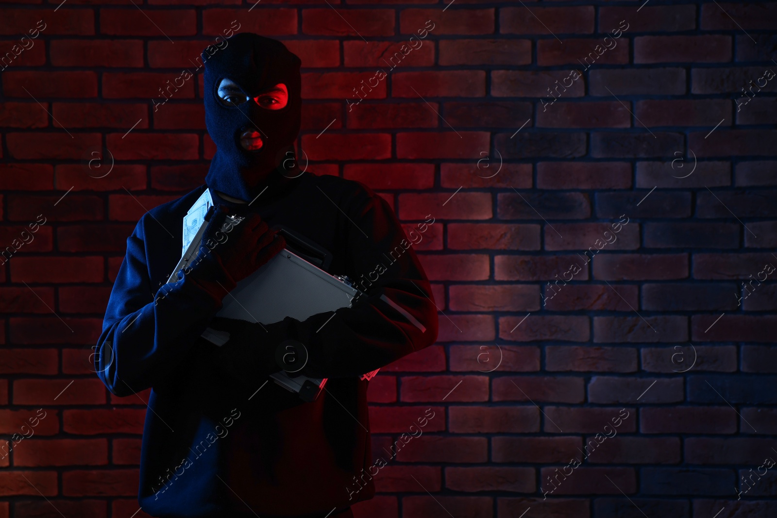 Photo of Thief in balaclava with briefcase of money in color lights against red brick wall. Space for text