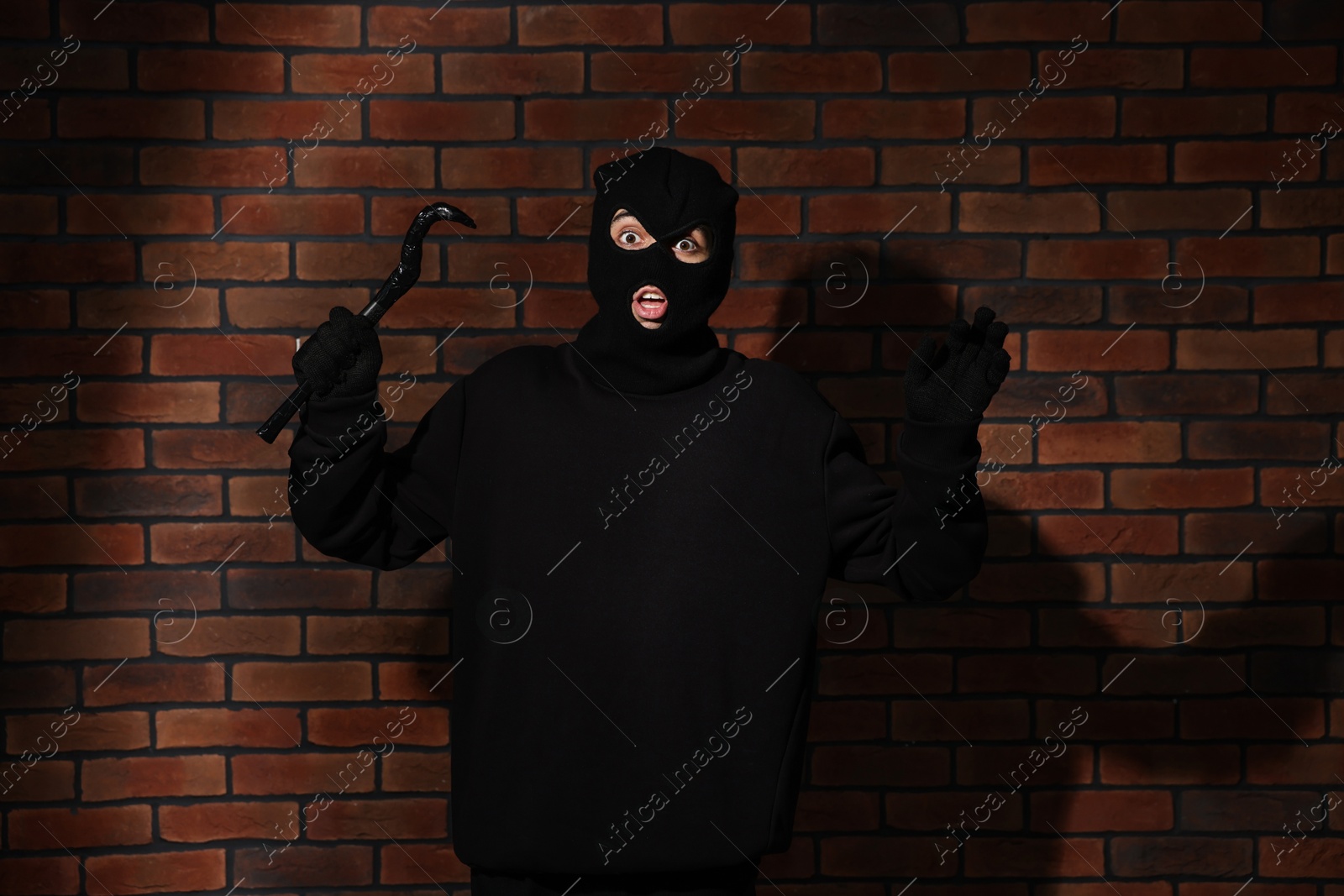 Photo of Emotional thief in balaclava raising hands with crowbar against red brick wall