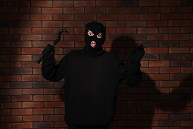 Photo of Emotional thief in balaclava raising hands with crowbar against red brick wall
