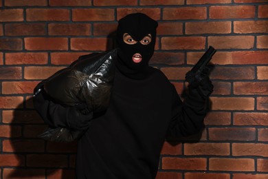 Photo of Emotional thief in balaclava with gun and bag against red brick wall