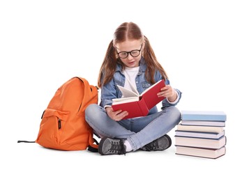 Cute little girl reading book near backpack on white background