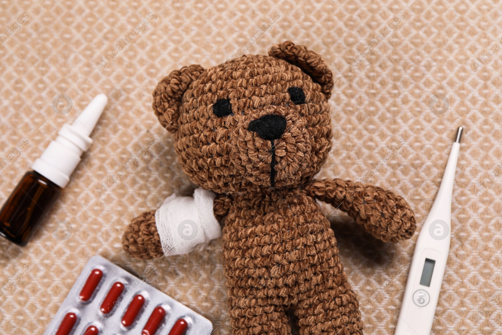 Photo of Toy bear with bandage, thermometer, pills and nasal spray on blanket, flat lay