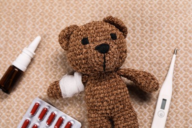 Toy bear with bandage, thermometer, pills and nasal spray on blanket, flat lay