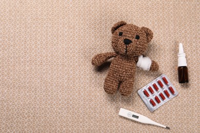 Toy bear with bandage, thermometer, pills and nasal spray on blanket, flat lay. Space for text