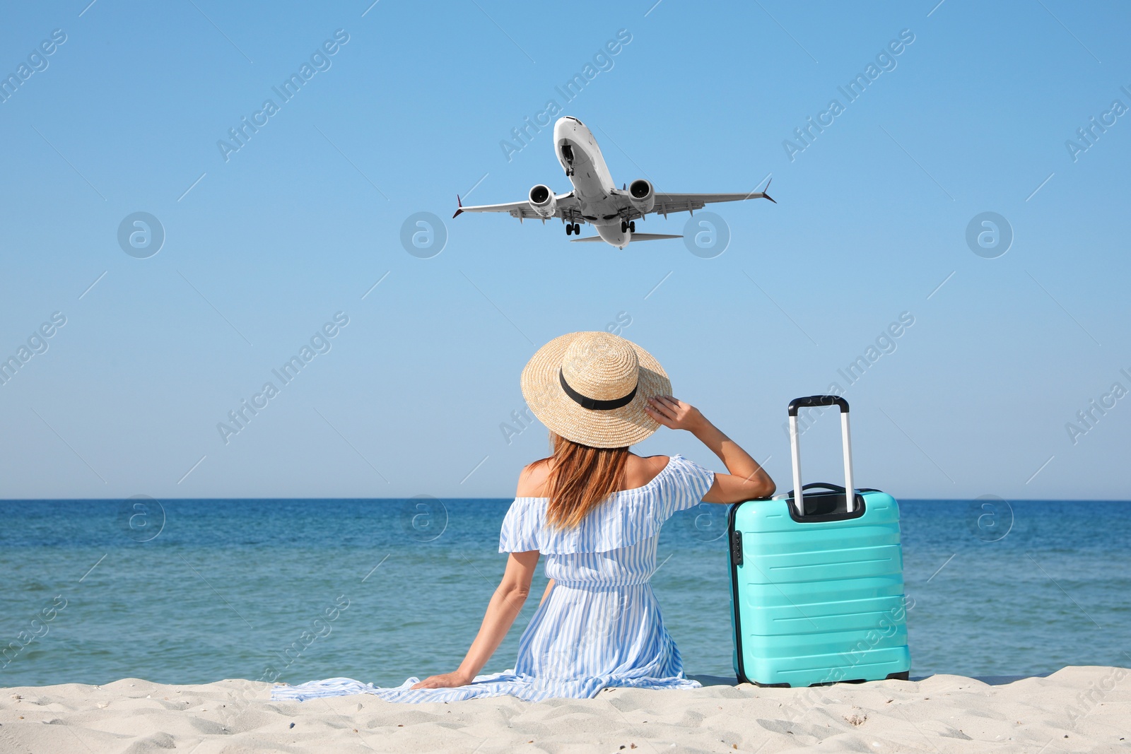 Image of Woman with suitcase on sandy beach looking at airplane flying in sky, back view