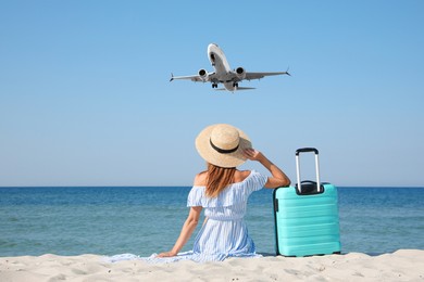 Woman with suitcase on sandy beach looking at airplane flying in sky, back view