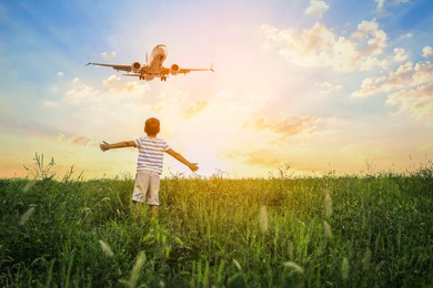 Boy in green meadow looking at airplane flying in sky during sunrise, back view