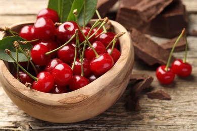 Photo of Fresh cherries with green leaves in bowl and dark chocolate on wooden table, closeup