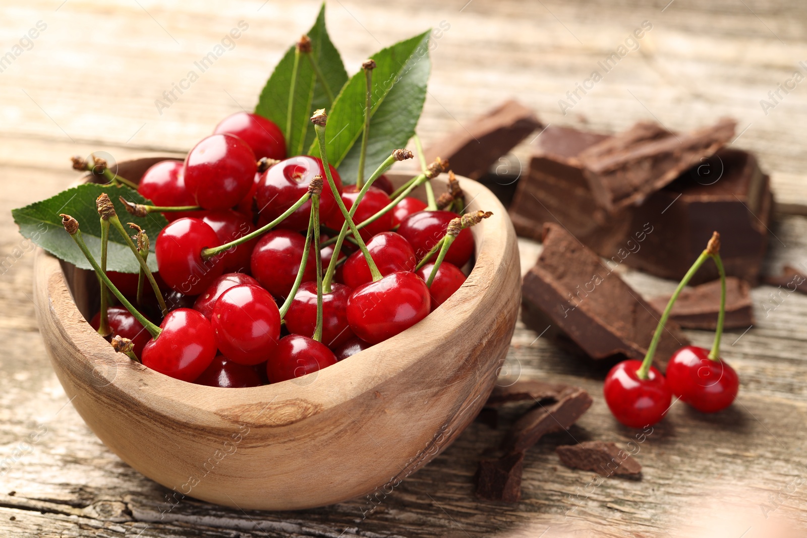 Photo of Fresh cherries with green leaves in bowl and dark chocolate on wooden table, closeup