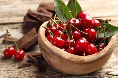 Fresh cherries with green leaves in bowl and dark chocolate on wooden table, closeup