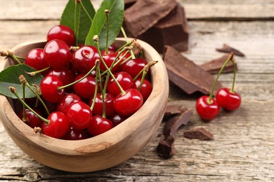 Photo of Fresh cherries with green leaves in bowl and dark chocolate on wooden table, closeup