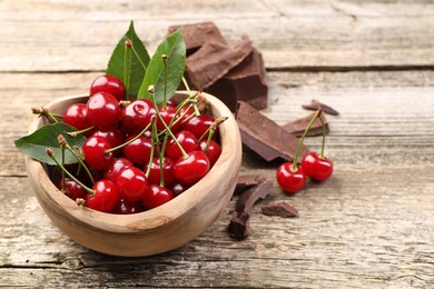 Fresh cherries with green leaves in bowl and dark chocolate on wooden table, space for text