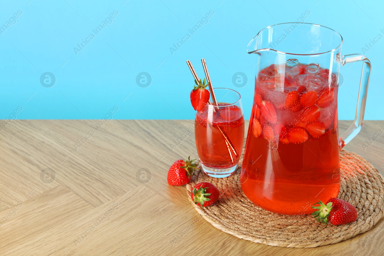 Photo of Tasty strawberry lemonade in jug, glass and berries on wooden table against light blue background. Space for text