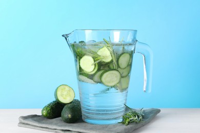 Refreshing cucumber water with rosemary in jug and vegetables on white table against light blue background