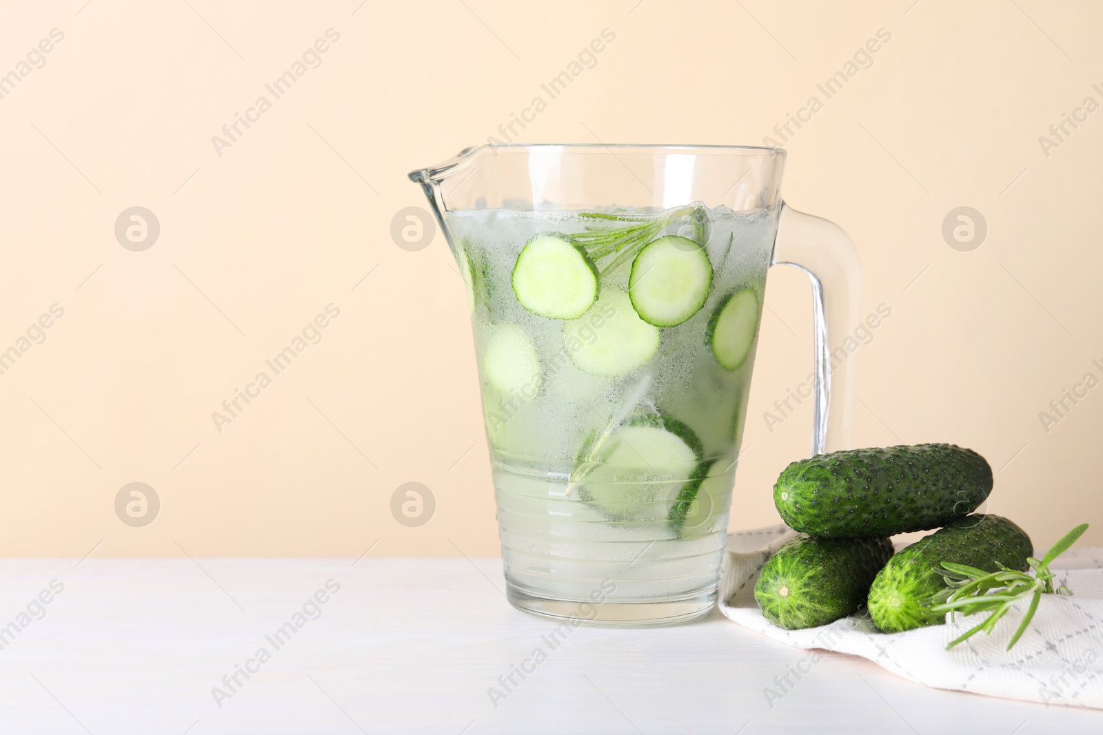 Photo of Refreshing cucumber water with rosemary in jug and vegetables on white table against beige background. Space for text