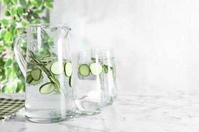 Refreshing cucumber water with rosemary in jug and glasses on white marble table. Space for text