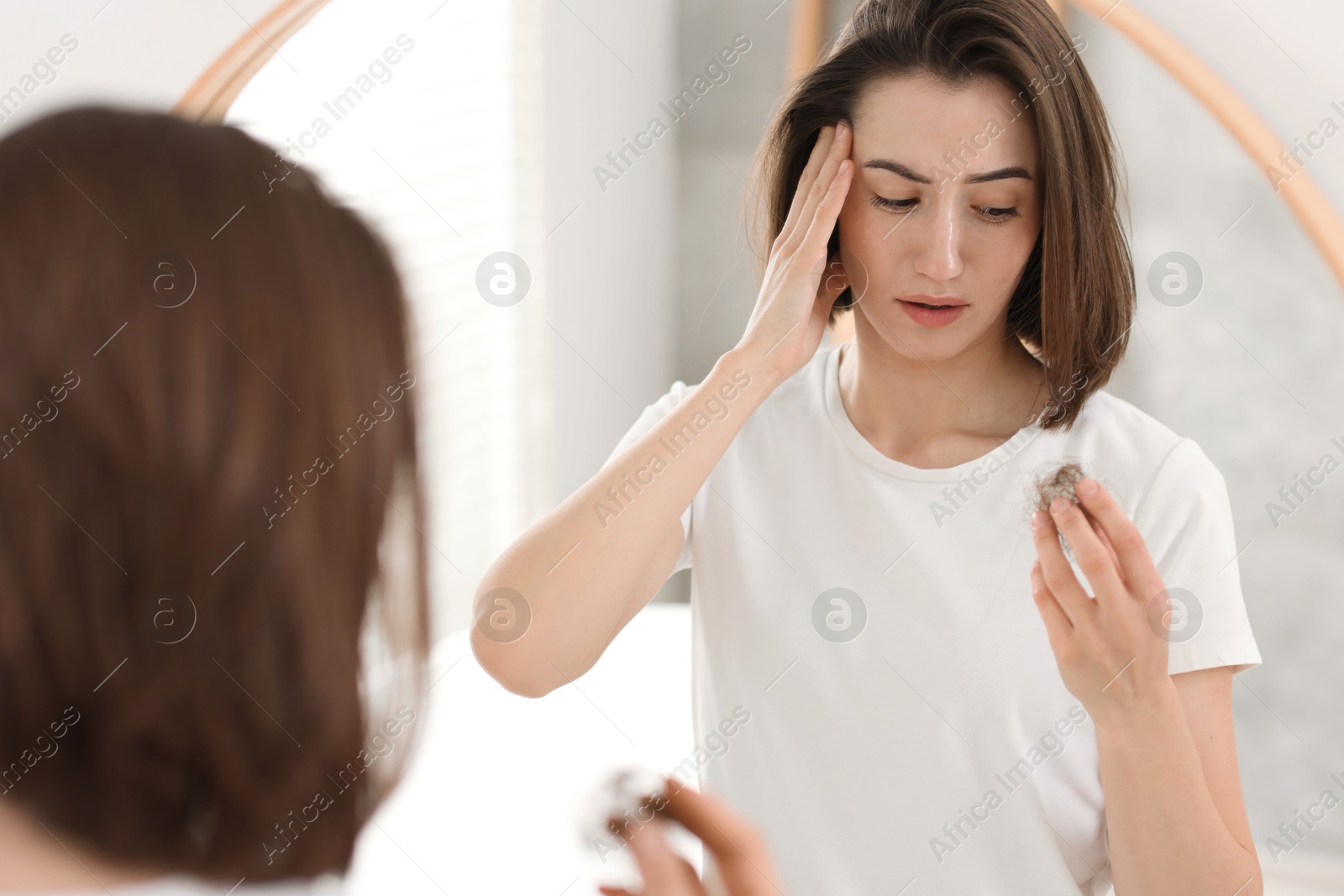 Photo of Stressed woman holding clump of lost hair near mirror indoors. Alopecia problem