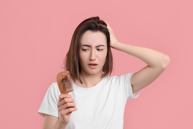 Sad woman holding comb with lost hair on pink background. Alopecia problem