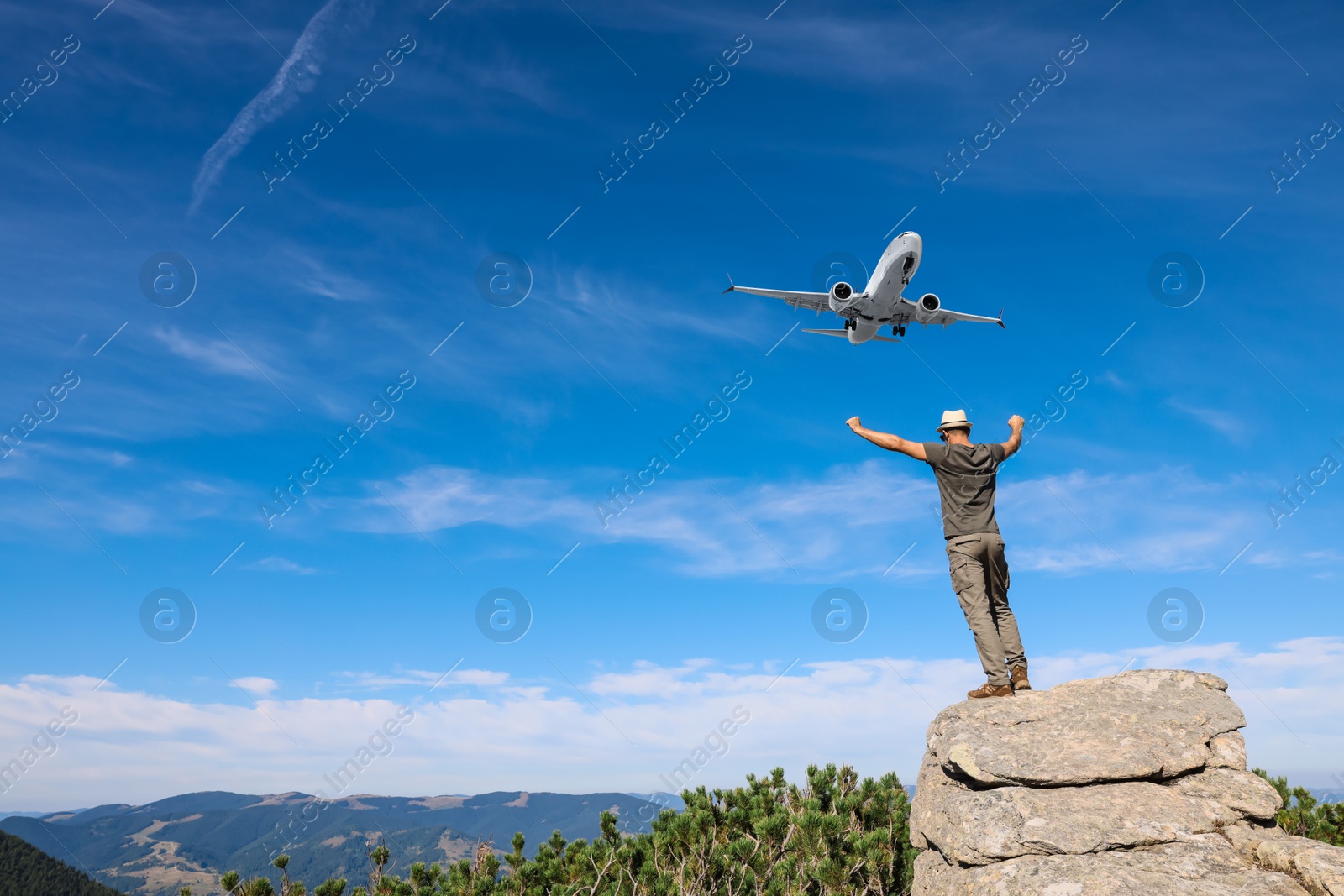 Image of Man on cliff looking at airplane flying in sky over mountains