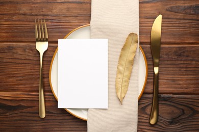 Photo of Empty menu, cutlery, plate, napkin and dry leaf on wooden table, top view. Mockup for design