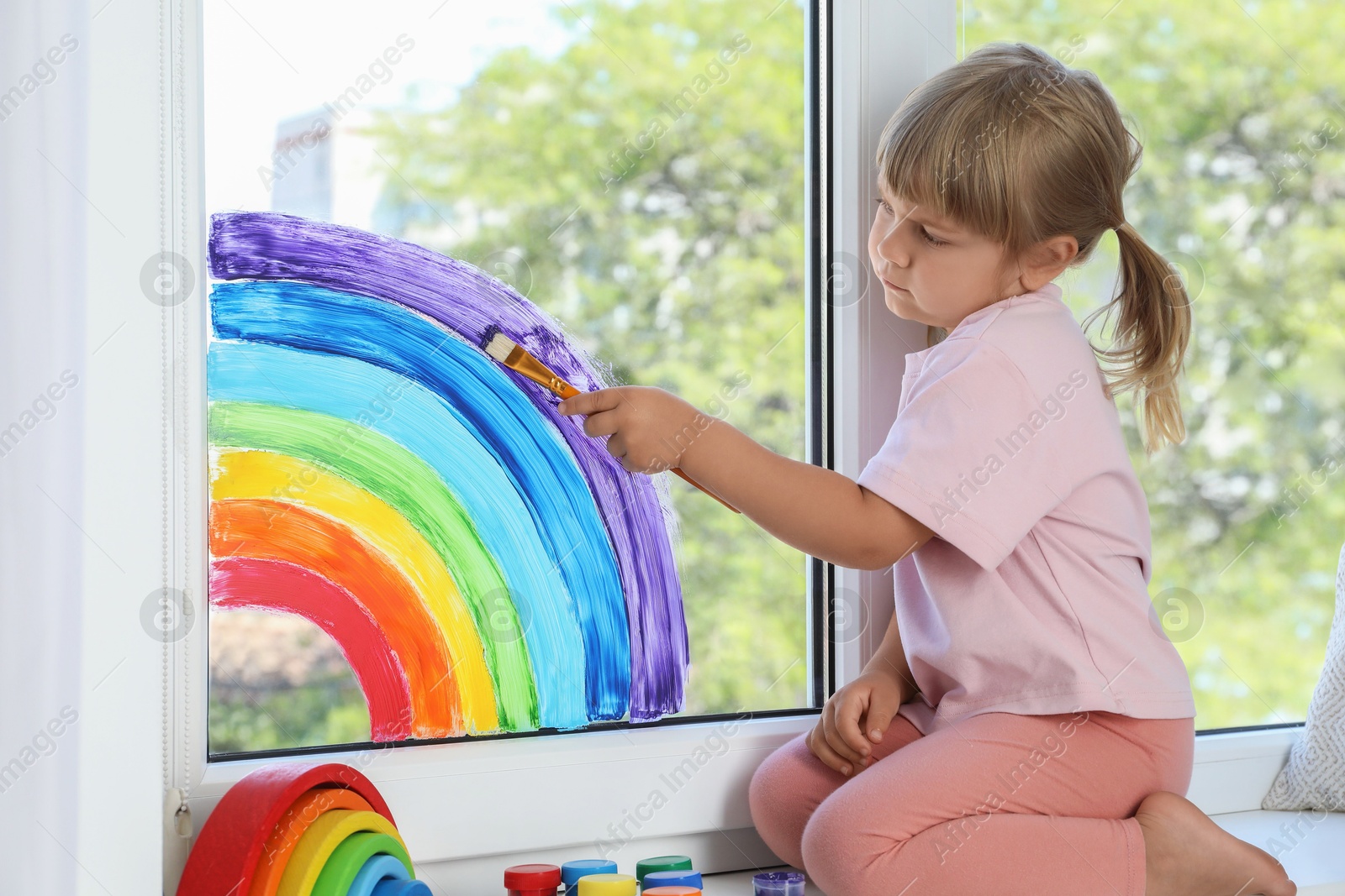 Photo of Little girl drawing rainbow on window indoors