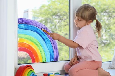 Little girl drawing rainbow on window indoors