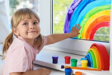 Photo of Little girl touching picture of rainbow on window indoors