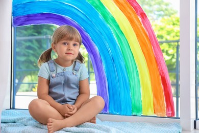 Photo of Little girl near rainbow painting on window indoors