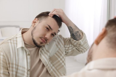 Photo of Baldness concept. Sad man with receding hairline looking at mirror indoors