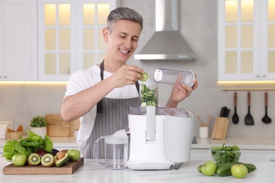 Photo of Smiling man putting fresh cucumber into juicer at white marble table in kitchen