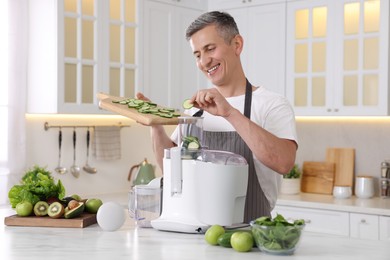 Photo of Smiling man putting fresh cucumber into juicer at white marble table in kitchen