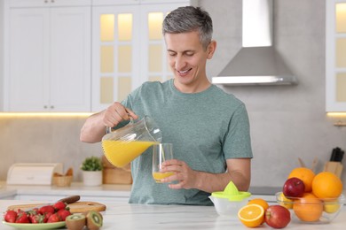 Smiling man pouring fresh orange juice into glass at white marble table in kitchen