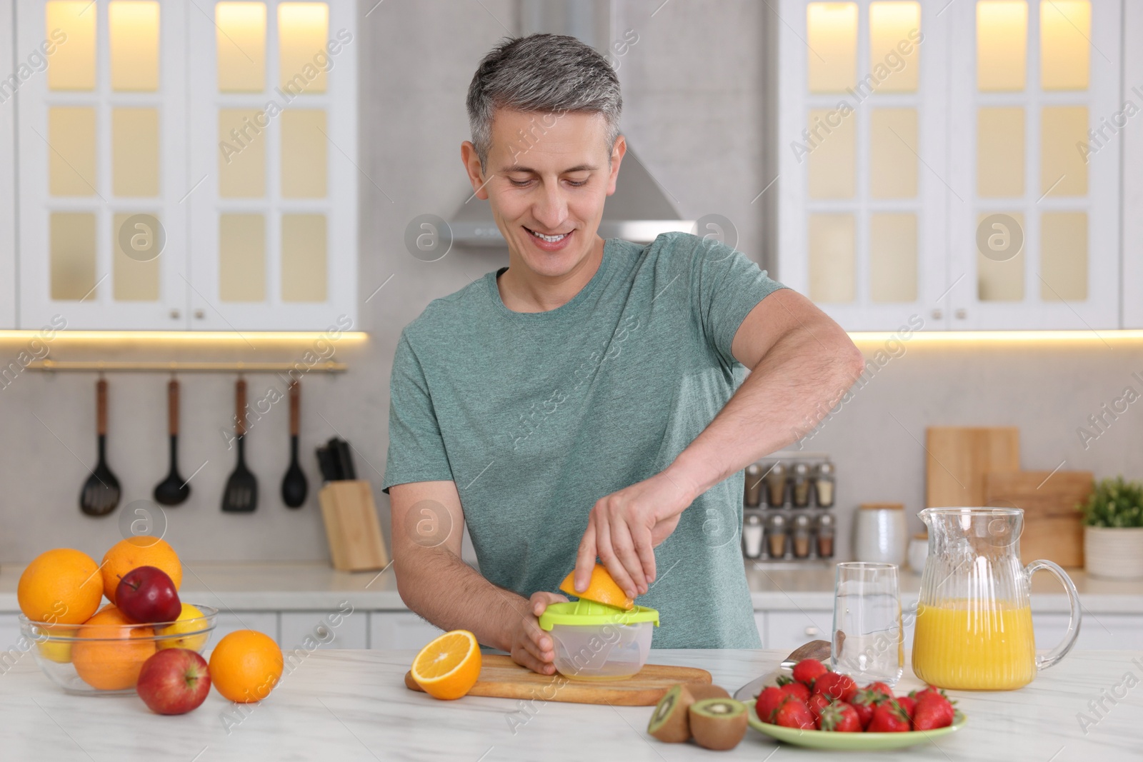 Photo of Smiling man squeezing fresh orange with juicer at white marble table in kitchen