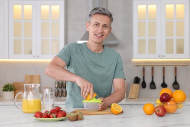 Smiling man squeezing fresh orange with juicer at white marble table in kitchen