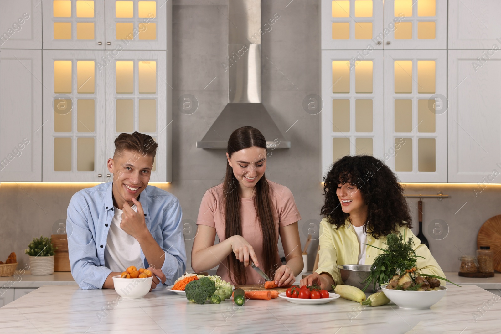 Photo of Friends cooking healthy vegetarian meal at white marble table in kitchen