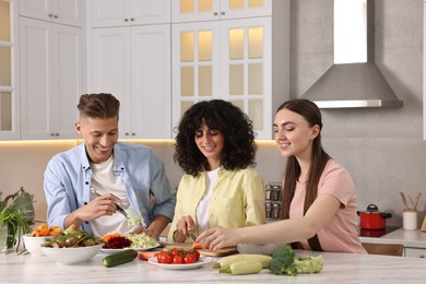 Photo of Friends cooking healthy vegetarian meal at white marble table in kitchen