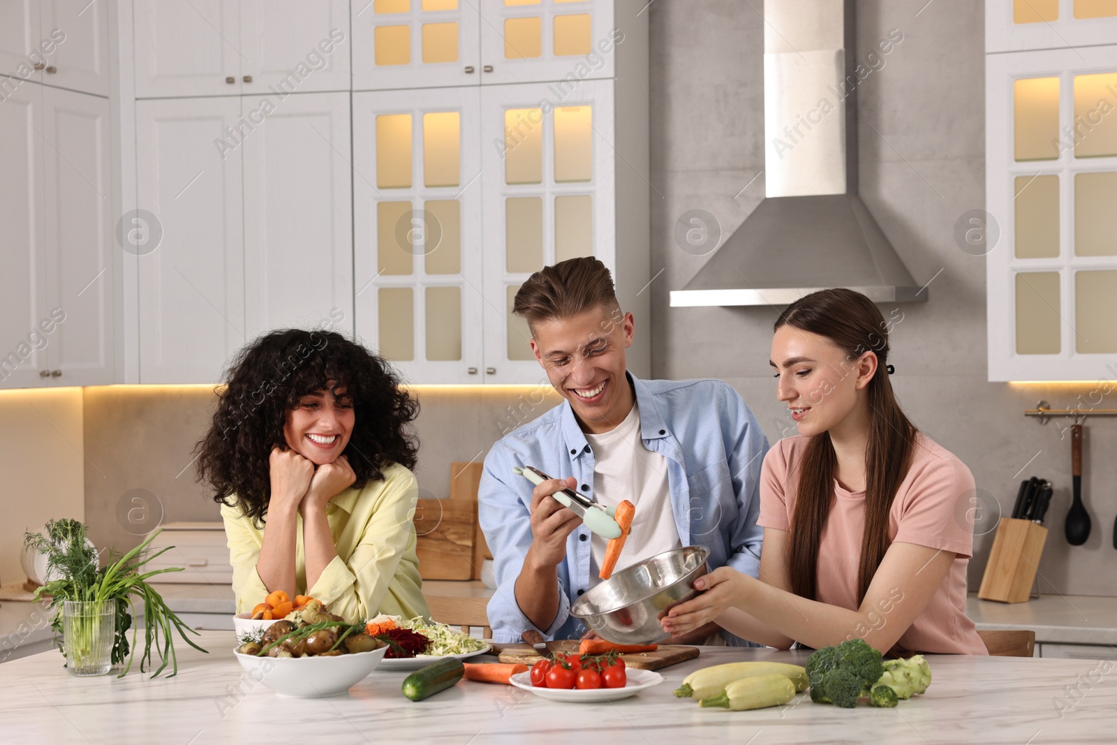 Photo of Friends cooking healthy vegetarian meal at white marble table in kitchen