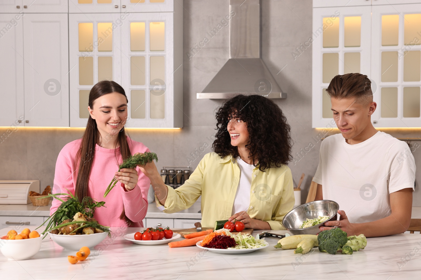 Photo of Friends cooking healthy vegetarian meal at white marble table in kitchen